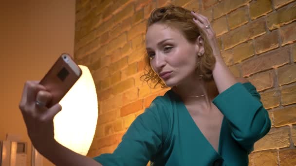 Close-up πορτρέτο του τζίντζερ σγουρά μαλλιά κορίτσι Καυκάσιος κάνοντας selfie φωτογραφίες αγγίζει τα μαλλιά χρησιμοποιώντας το smartphone στο φιλόξενο σπίτι φόντο. — Αρχείο Βίντεο