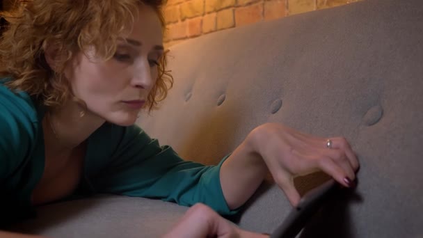 Close-up πορτρέτο στο προφίλ του τζίντζερ σγουρά μαλλιά κορίτσι Καυκάσιος ξαπλωμένος στον καναπέ και πληκτρολογώντας στο tablet σκεπτικά σχετικά άνετο σπίτι φόντο. — Αρχείο Βίντεο