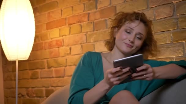 Retrato de jengibre de pelo rizado chica caucásica sentada en el sofá y mirando en la tableta sonriente en el acogedor fondo del hogar . — Vídeos de Stock