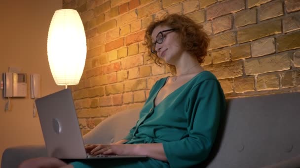 Primer plano retrato en el perfil de jengibre de pelo rizado chica caucásica en gafas de trabajo con el ordenador portátil sonriente en el acogedor fondo de la casa . — Vídeos de Stock