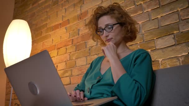 Ingwer lockige kaukasische Mädchen in Brille arbeiten mit Laptop nachdenklich und verschränken ihre Arme auf gemütlichen Hintergrund zu Hause. — Stockvideo