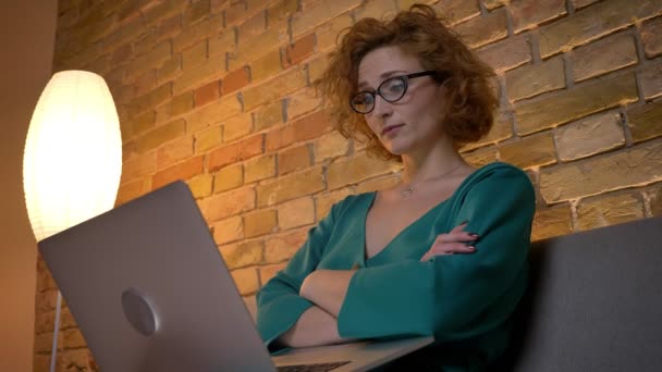 Ginger kręcone włosy kaukaski dziewczyna w okularach pracę z laptopem radośnie i broni jej przekraczania na przytulnym domu tło. — Wideo stockowe