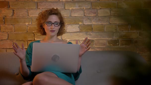 Retrato de gengibre de cabelos cacheados menina caucasiana em óculos sentados no sofá na frente do laptop mostra extrema surpresa no aconchegante casa fundo . — Vídeo de Stock