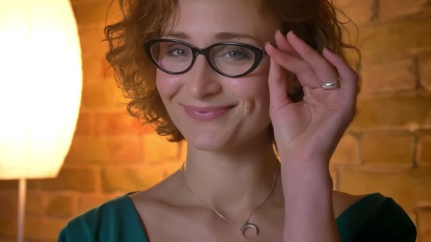 Nahaufnahme Porträt eines kaukasischen Mädchens mit lockigem Haar, das kokett seine Brille richtet und vor heimeligem Hintergrund in die Kamera lächelt. — Stockvideo
