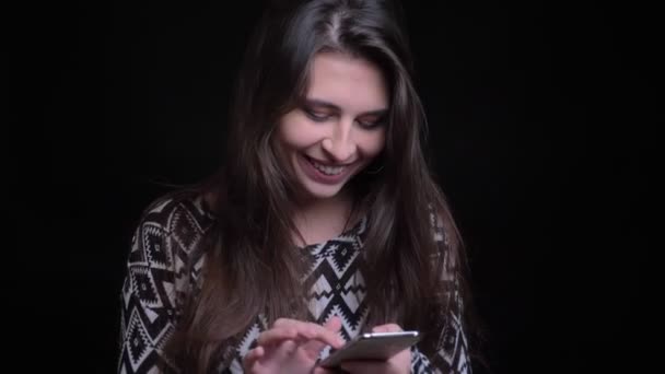 Крупный план портрета молодой красивой кавказской женщины, использующей телефон и смеющейся на фоне, изолированном от черного — стоковое видео