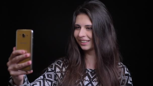 Closeup schieten van het jonge aantrekkelijke Kaukasische vrouwelijke hebben een video-oproep op de telefoon hallo zwaaien en lachend met geïsoleerd op zwarte achtergrond — Stockvideo