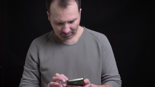 Portrait d'homme caucasien concentré d'âge moyen avec boucle d'oreille regardant sérieusement dans le smartphone sur fond noir . — Video