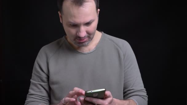 Portret van middelbare leeftijd geconcentreerd Kaukasische man met earring kijken vreugdevol in smartphone op zwarte achtergrond. — Stockvideo