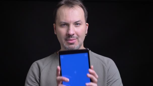 Middle-aged concentrated caucasian man with earring shows upright blue screen of tablet into camera on black background. — Stock Video