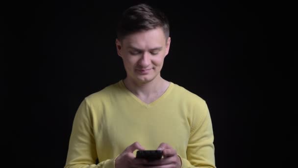 Retrato de hombre caucásico de mediana edad en suéter amarillo mirando sonriente en el teléfono inteligente y en la cámara sobre fondo negro . — Vídeo de stock