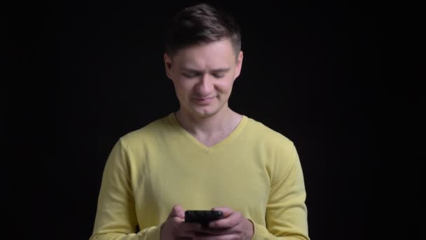 Портрет чоловіка середнього віку в жовтому светрі, який посміхається в смартфон на чорному тлі . — стокове відео