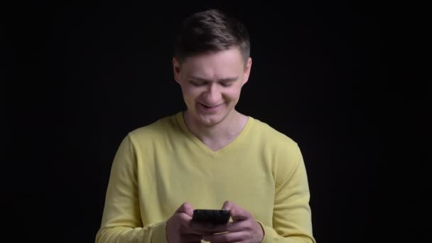 Retrato de hombre caucásico de mediana edad en suéter amarillo mirando en el teléfono inteligente y reaccionando emocionalmente sobre fondo negro . — Vídeo de stock