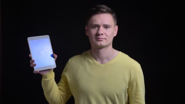 Middle-aged caucasian man in yellow sweater showing blue screen of tablet joyfully into camera on black background. — Stock Video