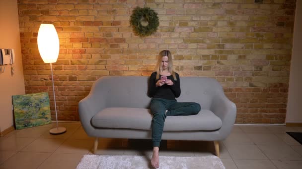 Closeup portrait of young pretty blonde-haired female using the phone and sitting on the couch indoors at cozy home — Stock Video