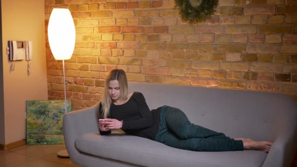 Крупним планом портрет молодої красивої блондинки, що використовує телефон і лежить на дивані, відпочиваючи в приміщенні в затишному будинку — стокове відео
