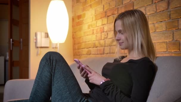 Портрет молодой симпатичной блондинки-студентки с телефоном, сидящей на диване, затем смотрящей в камеру и улыбающейся в уютном доме — стоковое видео
