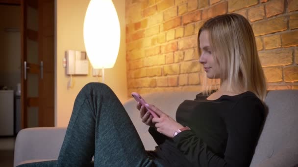 Zbliżenie po stronie portretu z młodych kobiet student siedząc na kanapie a następnie patrząc na kamery i uśmiechający się w pomieszczeniu o przytulny dom za pomocą telefonu — Wideo stockowe