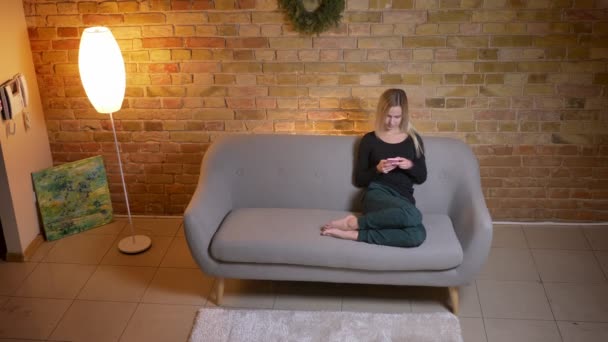 Närbild ovanifrån porträtt av unga ganska blonda kvinna använder telefonen och sitter i soffan inomhus på mysiga hem — Stockvideo