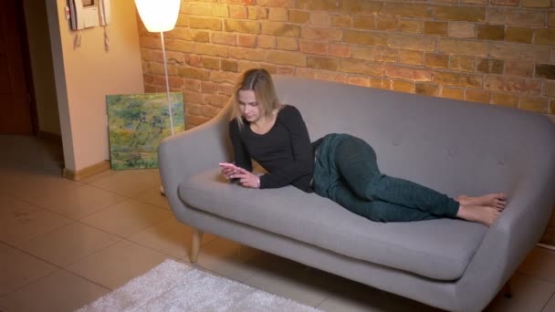 Primer plano superior disparar retrato de la joven mujer relajada utilizando el teléfono y acostado en el sofá descansando en el interior en casa acogedora — Vídeo de stock