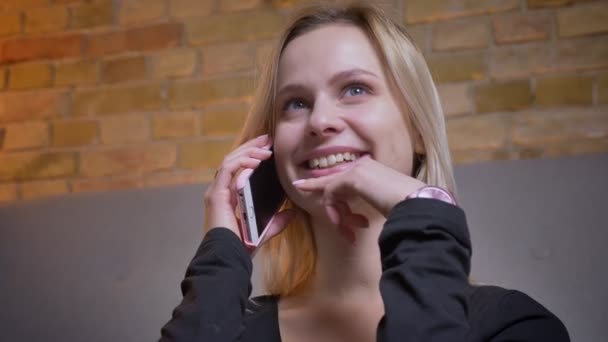 Retrato de cerca de una jovencita guapa de cabello rubio conversando por teléfono sonriendo y hablando emocionalmente en el interior de una casa acogedora — Vídeo de stock