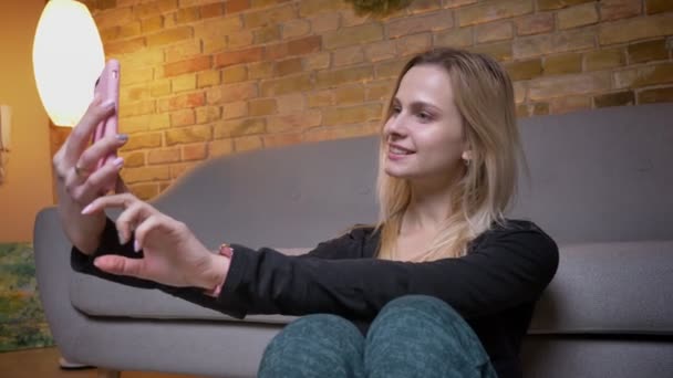 Closeup portret van jonge vrouwelijke student nemen van selfies op de telefoon en glimlachend zittend op de vloer leunend op de couch binnenshuis gezellig thuis — Stockvideo