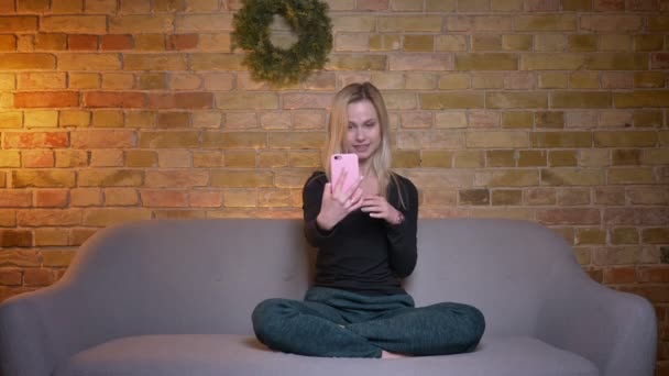 Крупный план портрета молодой милой девушки-подростка, которая разговаривает по телефону и улыбается, сидя на диване в уютном доме — стоковое видео