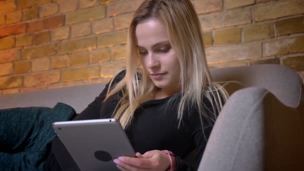 Portrait rapproché d'une jeune jolie femme aux cheveux blonds utilisant la tablette et allongée sur le canapé allongée à l'intérieur dans une maison confortable — Video