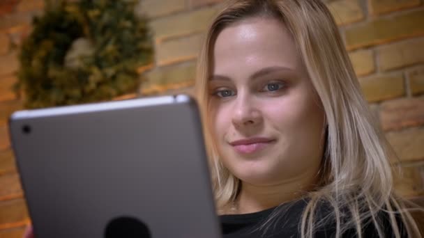 Närbild porträtt av ung kvinna med blont hår använder tabletten inomhus mysiga hemma — Stockvideo