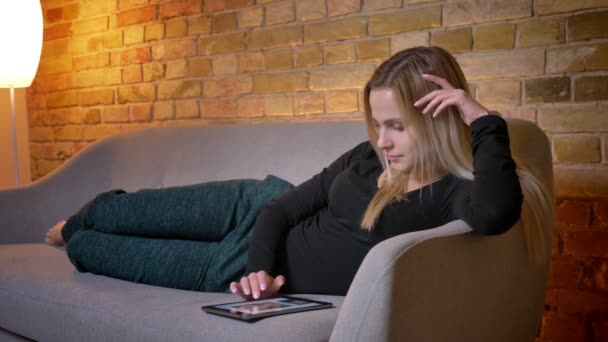 Nahaufnahme Porträt einer jungen hübschen Hausfrau, die das Tablet benutzt und zu Hause entspannt auf dem Sofa liegt — Stockvideo