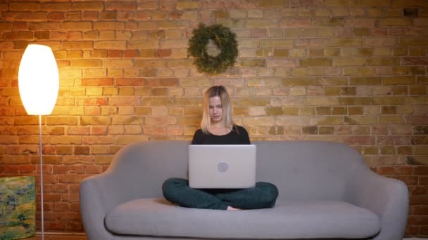 Retrato de cerca de una joven guapa rubia usando el portátil y sentada en el sofá dentro de una casa acogedora — Vídeos de Stock