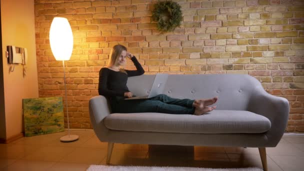 Retrato de vista lateral de jovem linda millennial feminino assistindo uma comédia no laptop e sorrindo alegremente enquanto sentado no sofá dentro de casa aconchegante — Vídeo de Stock