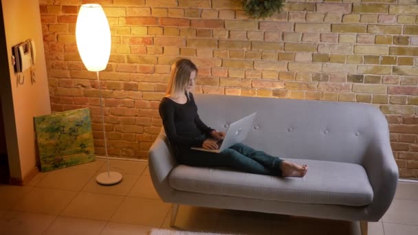 Retrato de vista lateral de una joven estudiante bastante femenina que estudia en línea en el portátil mientras está sentada en el sofá en el interior de una casa acogedora — Vídeos de Stock