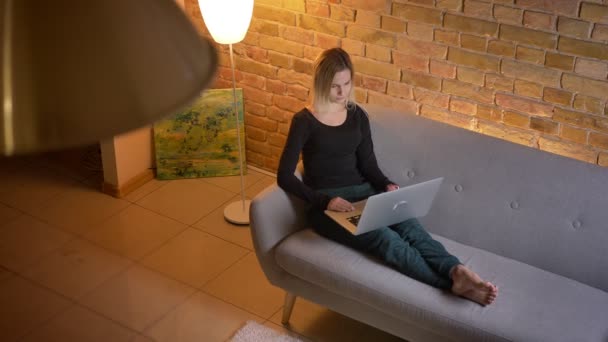 Стрілянина молодої красивої бізнес-леді, що працює дистанційно на ноутбуці, сидячи на дивані в приміщенні в затишному будинку — стокове відео