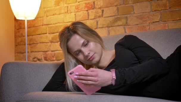 Retrato de cerca de una joven guapa rubia usando el teléfono y tumbada en el sofá descansando, luego mirando a la cámara y sonriendo en el interior de una acogedora casa — Vídeo de stock
