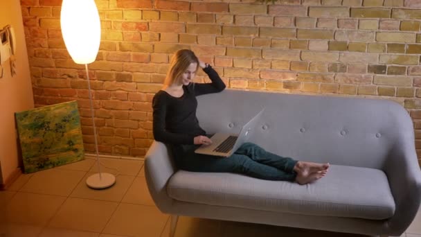 Portrait de jeune jolie femme au foyer regardant un film sur l'ordinateur portable et souriant joyeusement tout en étant assis sur le canapé à l'intérieur dans un appartement confortable — Video