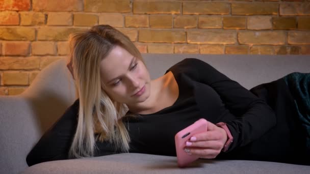 Närbild porträtt av ung ganska blont hemmafru använder telefonen och liggande på soffan lutar på hennes hand inomhus på mysiga hem. — Stockvideo