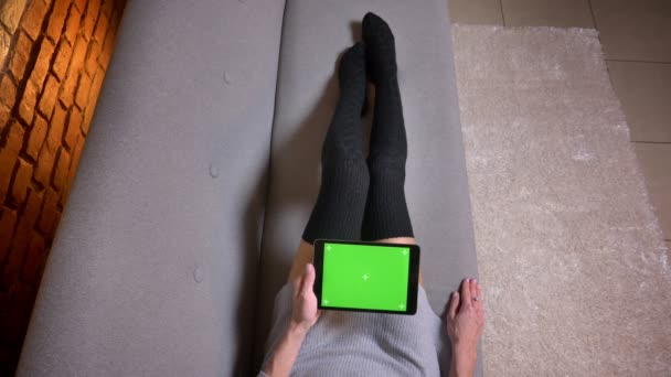 Крупный план съемки женских ног в милых носках на диване в помещении. Женщина просматривает планшет с зеленым экраном — стоковое видео
