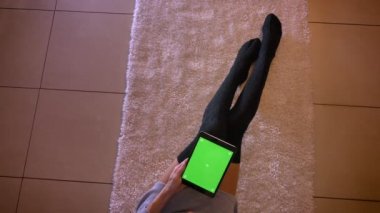 Portre sevimli genç kız bacaklarını kapalı halı üzerinde şirin çoraplı portresi. Kadın katta oturan ve tablet cihazın dikey tutarak yeşil ekranda kaydırma