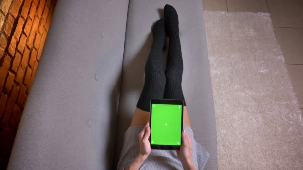 Closeup tiro de pernas femininas em meias bonitos no sofá dentro de casa. Mulher segurando o comprimido com tela verde verticalmente — Vídeo de Stock