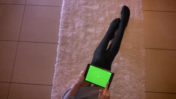 Portrait rapproché de jolies jambes d'adolescentes en chaussettes mignonnes sur le tapis à l'intérieur. Femme assise sur le sol et faisant défiler la tablette sur l'écran vert — Video