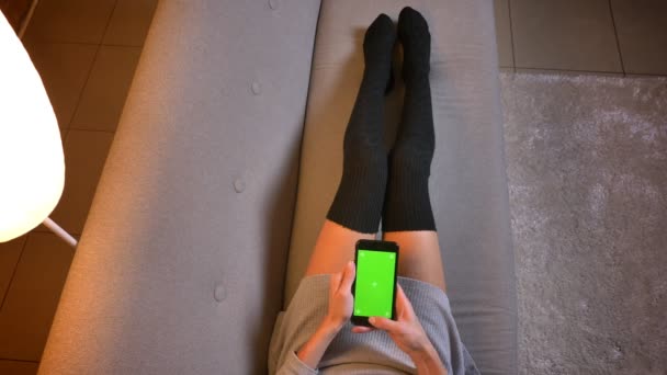 Vértes lő a fiatal, vonzó női szociális média influencer a telefon használata a zöld képernyő. Womans lábak aranyos zokni a kanapén — Stock videók