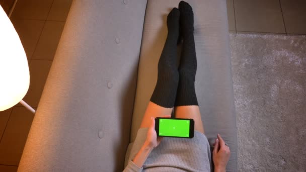 Closeup střílet z mladé dospívající žena drží telefon s zelenou obrazovkou s reklamou na něm. Zenske nohy v roztomilé ponožky na gauči — Stock video