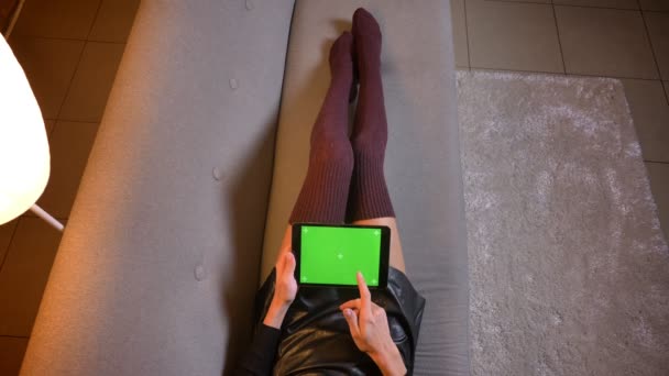 Gros plan shoot of female social media influenceur shopping en ligne sur la tablette avec écran chromatique vert. Cuisse femme en mignon tricot chaussettes et jupe en cuir sur le canapé — Video