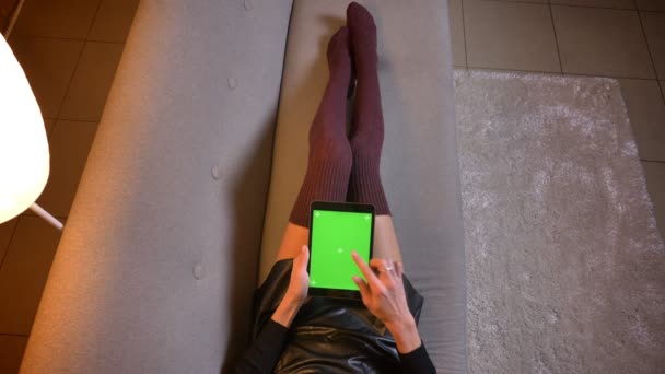 Nahaufnahme einer jungen Frau, die ein Video auf dem Tablet mit grünem Chrombildschirm anschaut. Frauenschenkel in süßen Stricksocken und Lederrock auf der Couch — Stockvideo