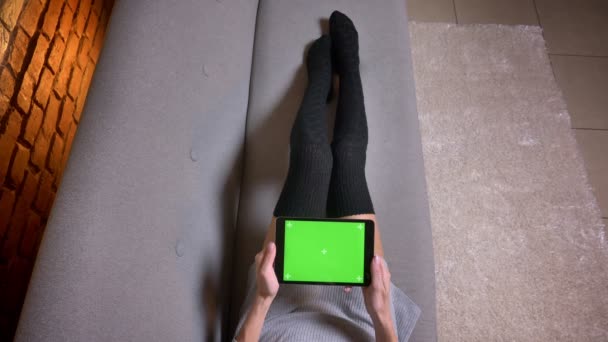 Крупним планом знімають жіночі ноги в милих шкарпетках на дивані в приміщенні. Жінка тримає планшет з зеленим екраном горизонтально — стокове відео