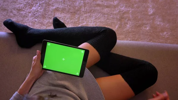 Closeup portrait of female beauty blogger using social media on the tablet with green screen. Girls legs in a cozy position in socks on the couch