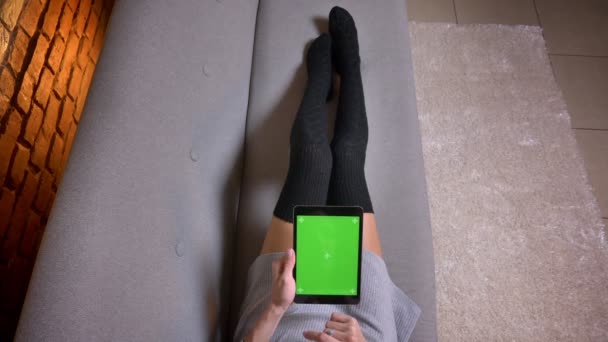 Nahaufnahme von jungen Mädchen Beine in süßen Socken auf dem Sofa drinnen. zarte Frau hält das Tablet mit grünem Bildschirm horizontal und scrollt