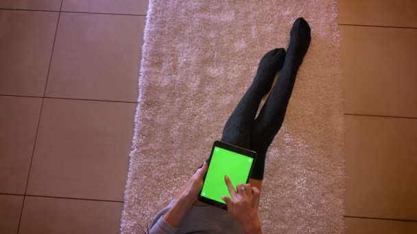 Portrait rapproché de jolies jambes d'adolescentes en chaussettes mignonnes sur le tapis à l'intérieur. Femme assise sur le sol et regardant une vidéo sur la tablette sur l'écran vert — Video