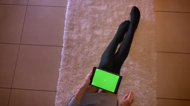 Portrait rapproché de jolies jambes d'adolescentes en chaussettes mignonnes sur le tapis à l'intérieur. Femme assise sur le sol et tenant la tablette avec l'écran vert horizontalement — Video