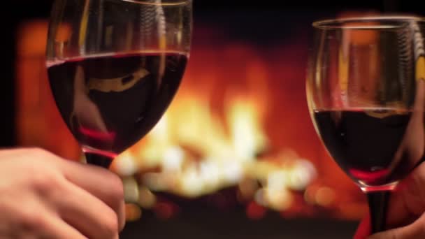 Gros plan pousse de deux mains d'amis cliquetis verres avec du vin rouge célébrant avec cheminée chaleureuse confortable sur le fond à l'intérieur dans la soirée — Video